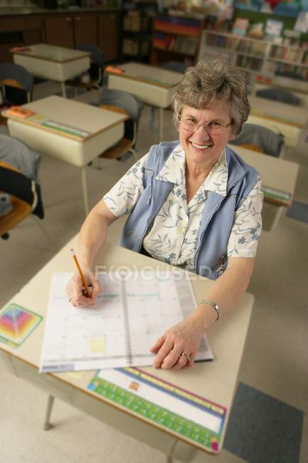 Feliz madura profesora sentada en el escritorio de la escuela - foto de stock