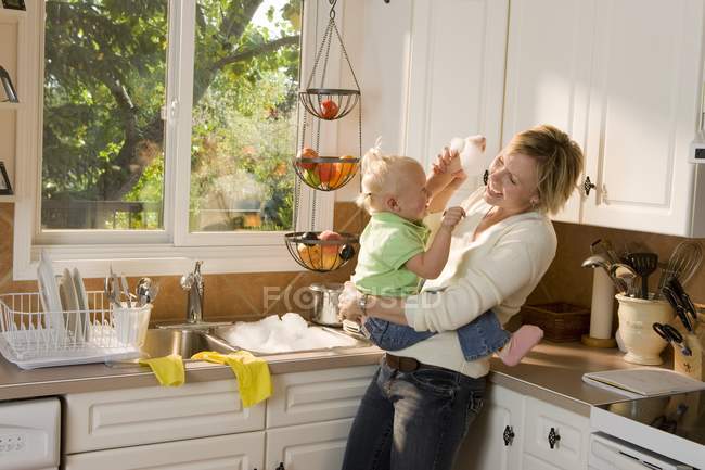 Madre caucásica e hija bebé abrazándose en la cocina y divirtiéndose - foto de stock