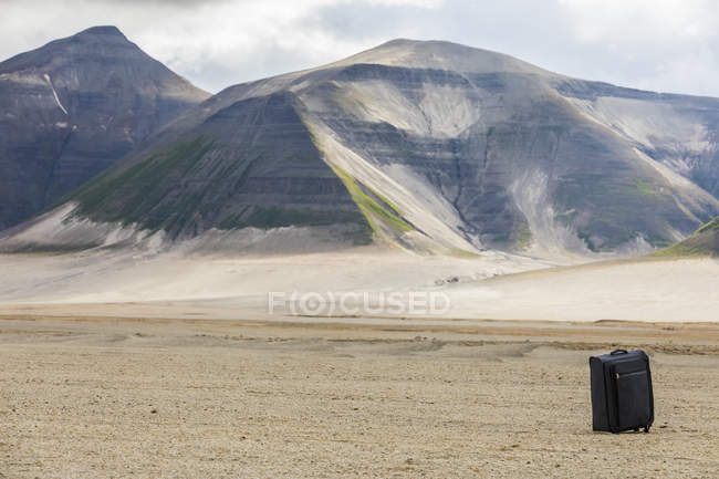 Vista do campo arenoso com colinas rochosas sob céu nublado no fundo e mala preta no chão durante o dia — Fotografia de Stock