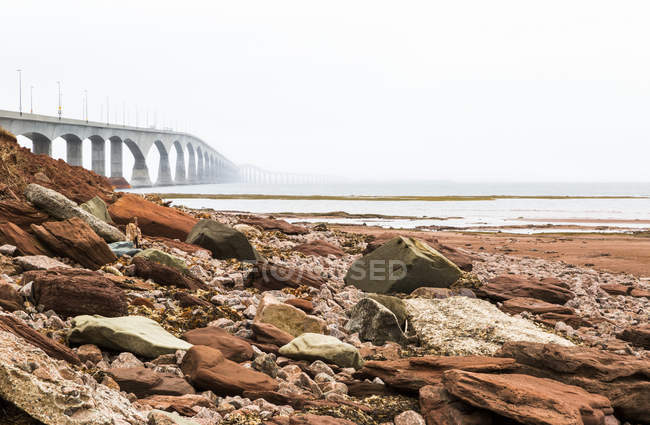 Sandy beach with pile of stones against water and bridge over water — Stock Photo