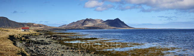 Paysage le long de la côte avec des pics de montagne au loin et un petit bâtiment avec un toit rouge le long du bord de l'eau, péninsule Snaefellsnes ; Islande — Photo de stock