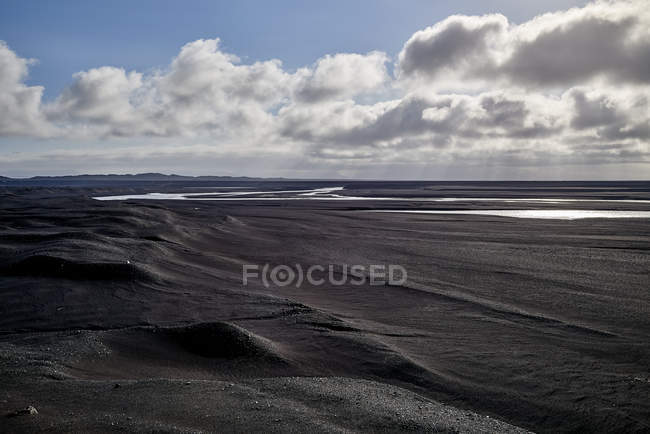 Ein Fluss, der durch ein Flussbett aus schwarzem Sand fließt, mit Bergen in der Ferne; Island — Stockfoto