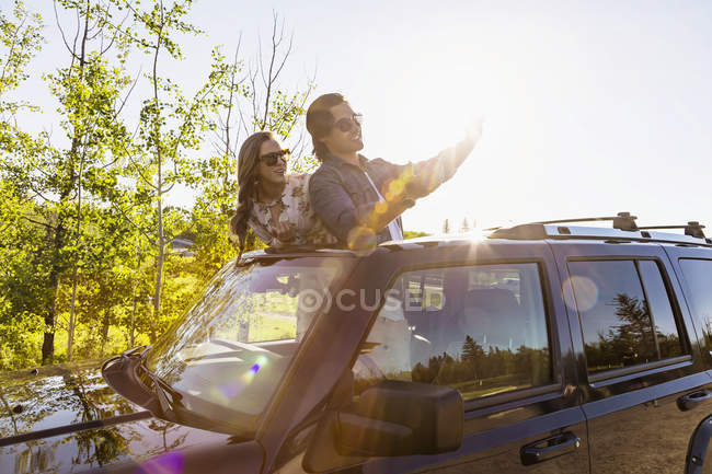 Молодая пара стоит у машины с откидным верхом и делает селфи на фоне деревьев — стоковое фото