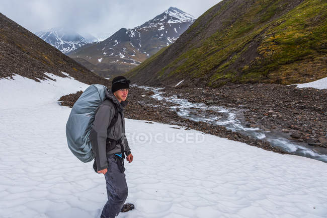 Homem com mochila coberta em pé sobre o campo de montanha com neve e picos no fundo — Fotografia de Stock
