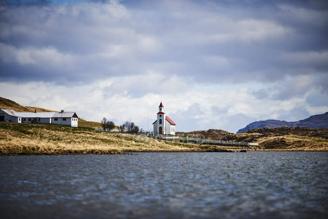 Iglesia con un tejado rojo, granero y granja a lo largo de un lago; Islandia - foto de stock