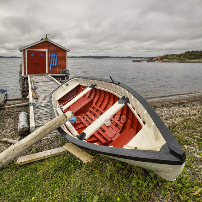Невеликий дерев'яний червоний сарай над водою на пірсі з дерев'яним човном і пагорбами на фоні — стокове фото