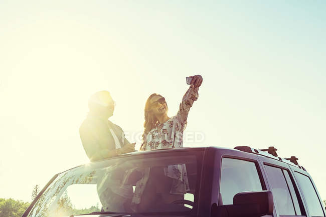 Молодая пара стоит у машины с откидным верхом и делает селфи на фоне солнечного света — стоковое фото