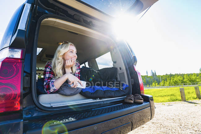 Блондинка молода дівчина з сонцезахисними окулярами лежить в машині і посміхається, дивлячись далеко — стокове фото