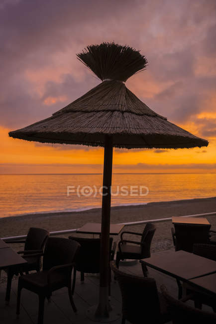 Un paraguas de paja sobre una mesa con sillas en la playa al atardecer, con vistas al mar Mediterráneo; Menton, Costa Azul, Francia - foto de stock
