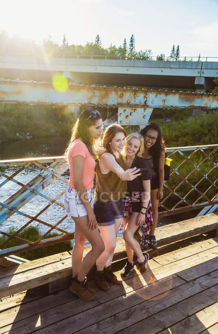 Четыре счастливые девушки стоят на деревянном балконе против металлического ограждения над водой и делают селфи — стоковое фото