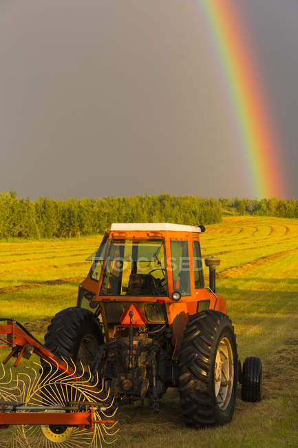Вид трактора, работающего на поле с инструментом и радугой на заднем плане в дневное время — стоковое фото