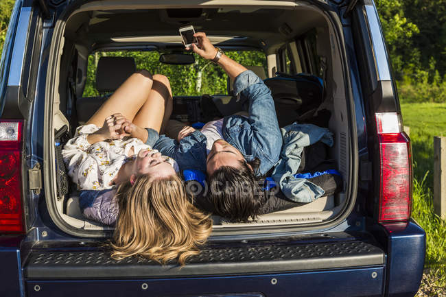 Junges Paar liegt am Auto und schaut tagsüber ins Smartphone und lächelt — Stockfoto