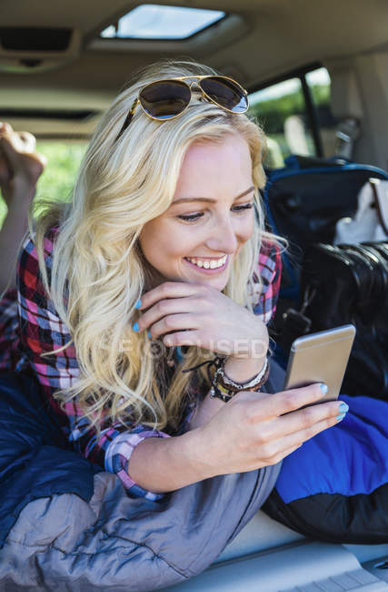 Glückliches Mädchen mit Sonnenbrille, das am Auto liegt und ins Smartphone schaut — Stockfoto