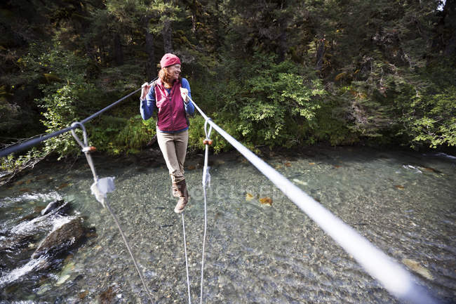 Una mujer camina en un cable en un puente colgante sobre un río; Alaska, Estados Unidos de América - foto de stock