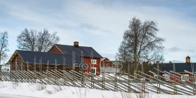 A Fence Lines A Farmyard With Red Farm Buildings In Winter; Arjeplog, Condado de Norrbotten, Suecia - foto de stock