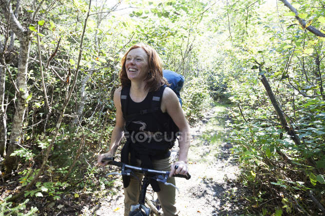 Una mujer se para en un camino en un bosque con su bicicleta y una mochila; Alaska, Estados Unidos de América - foto de stock