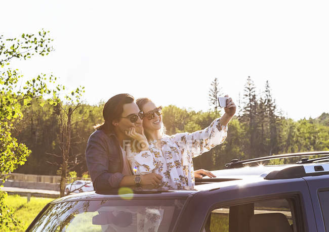 Yound pareja de pie en el coche con tapa de solapa y hacer selfie en el teléfono inteligente - foto de stock