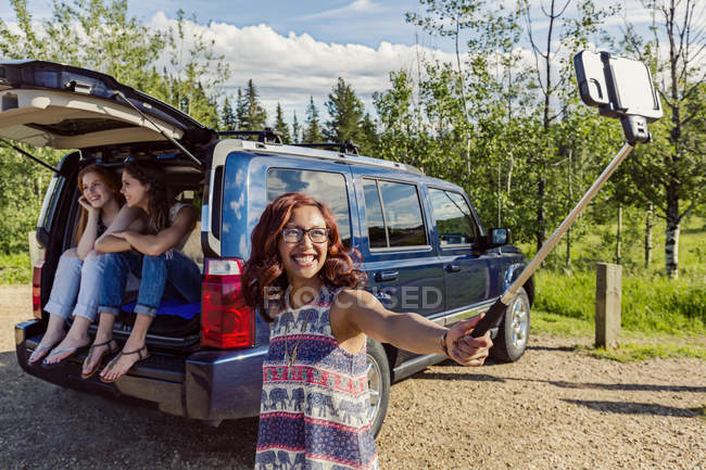 Three girls having fun, while two sitting at car another one making selfie by smartphone on monopod — Stock Photo