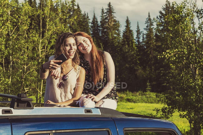 Duas meninas sentadas no topo do carro e abraçando uns aos outros com árvores no fundo — Fotografia de Stock