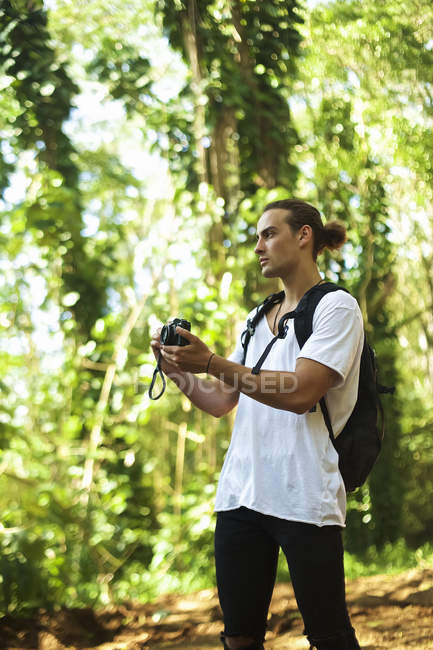 A young man stands on a trail wearing a backpack and taking a picture with a camera while on a hike; Hawaii, United States of America — Stock Photo