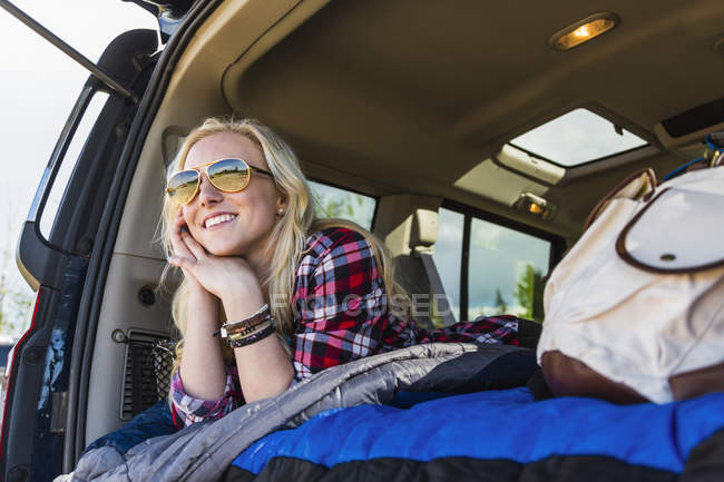 Blonde jeune fille en lunettes de soleil couché dans la voiture et souriant tout en regardant loin — Photo de stock