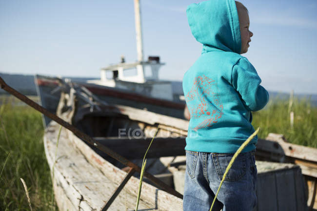 Un giovane ragazzo si trova accanto a una barca di legno sulla riva erbosa guardando verso l'acqua, Homer Spit; Homer, Alaska, Stati Uniti d'America — Foto stock