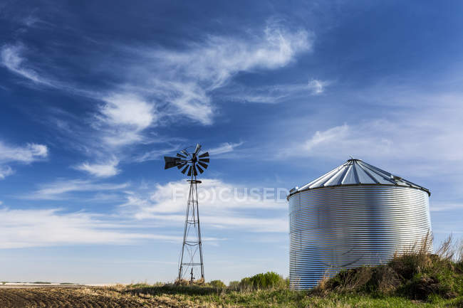 Un vecchio mulino a vento con un grande contenitore di grano metallico con interessanti nuvole e cielo blu; Beiseker, Alberta, Canada — Foto stock