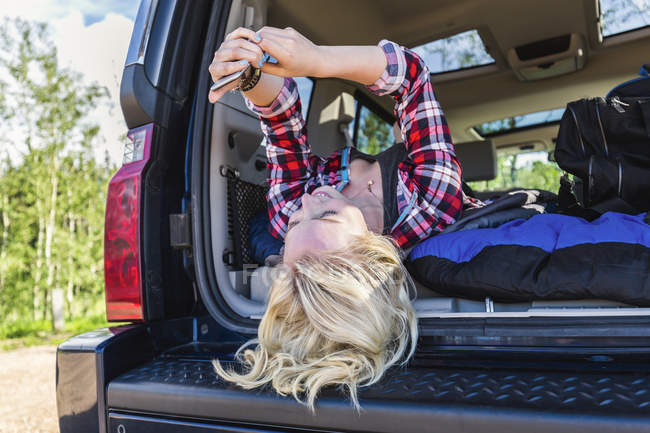 Chica rubia feliz tumbado en el coche y mirando el teléfono inteligente durante el día - foto de stock