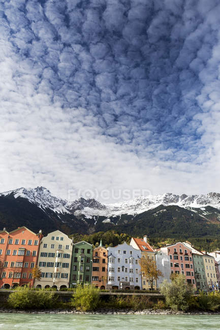 Edifícios coloridos ao longo da margem do rio com picos montanhosos cobertos de neve, nuvens dramáticas e céu azul por cima; Innsbruck, Tirol, Áustria — Fotografia de Stock