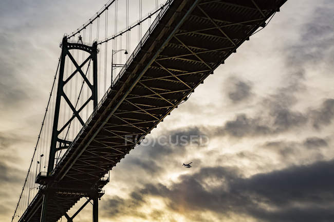 Vista de bajo ángulo de un puente al atardecer con un avión volando en la distancia; Vancouver, Columbia Británica, Canadá - foto de stock