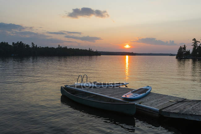 Le coucher du soleil sur un lac tranquille avec un quai, un canot et des planches à pagaie au premier plan ; Lac des Bois, Ontario, Canada — Photo de stock