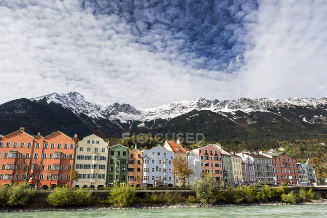 Edificios coloridos a lo largo de la orilla del río con picos de montaña cubiertos de nieve, nubes dramáticas y cielo azul; Innsbruck, Tirol, Austria - foto de stock