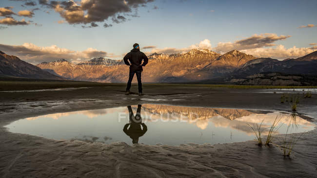 Hombre de pie con reflejo en una piscina de agua con vistas a las montañas de San Elías al atardecer, Parque Nacional y Reserva de Kluane, Destruction Bay, Yukón, Canadá - foto de stock