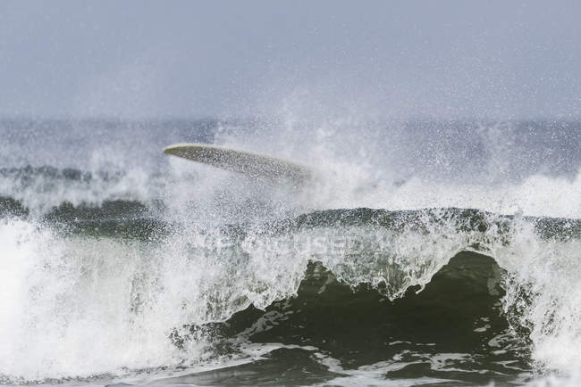 Olas rompiendo y estrellándose cerca de la costa con una tabla de surf en el agua salpicando en la parte superior de la ola; Tofino, Columbia Británica, Canadá - foto de stock