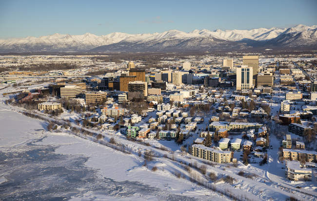 Aerial view of snow covering the sea ice on the frozen shores of downtown Anchorage, the Chugach Mountains in the distance beyond the office buildings and hotels, Cook Inlet in the foreground, South-central Alaska in winter; Anchorage, Alaska — Stock Photo