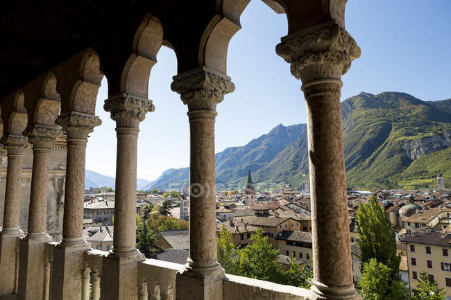 Le colonne di pietra di Palazzo incorniciano un villaggio alpino sullo sfondo con montagne e cielo azzurro; Trento, Trento, Italia — Foto stock