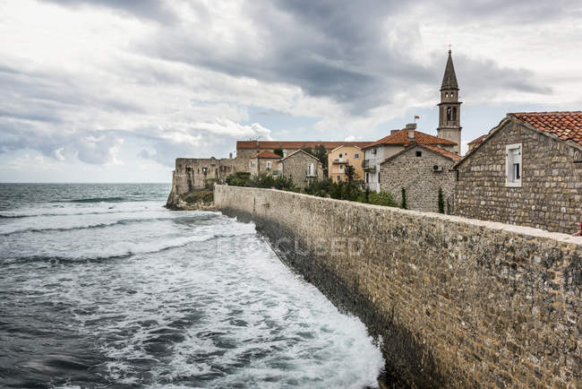Eine Steinmauer, die die Küste mit Gebäuden und einem Kirchturm säumt; budva, opstina budva, montenegro — Stockfoto