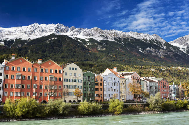 Colorata fila di edifici lungo una riva del fiume con catena montuosa innevata sullo sfondo e cielo azzurro; Innsbruck, Tirolo, Austria — Foto stock