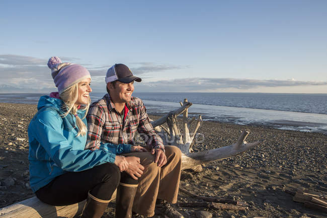 Un jeune couple est assis sur un morceau de bois flotté sur une plage donnant sur l'océan au coucher du soleil ; Anchorage, Alaska, États-Unis d'Amérique — Photo de stock