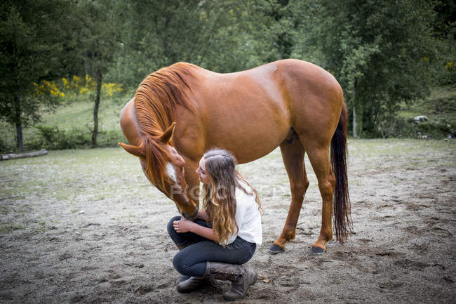 Una adolescente agachándose para hablar y cuidar de su caballo; Columbia Británica, Canadá - foto de stock