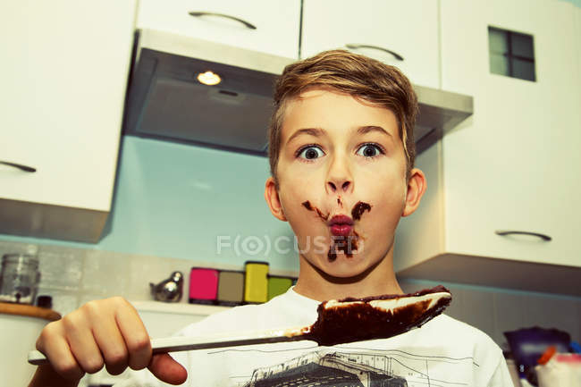 Giovane ragazzo con un viso disordinato con cioccolato dalla spatola dopo aver fatto fudge — Foto stock