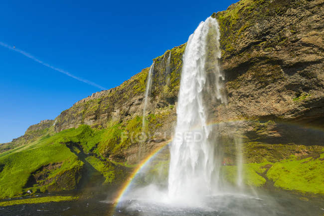 Cascada de Skogafoss con cielo azul y un arco iris en la niebla; Islandia - foto de stock