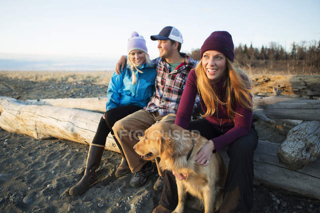 A young couple and a friend with a dog sit on a piece of driftwood on a beach looking out to the ocean at sunset; Anchorage, Alaska, United States of America — Stock Photo