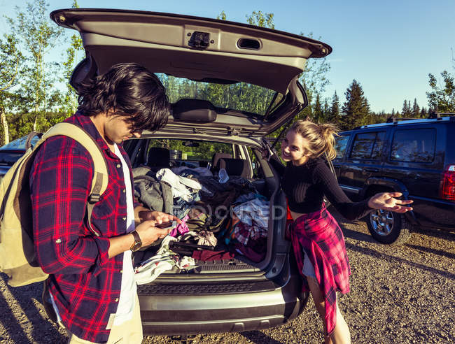 Молода пара стоїть в упакованому автомобілі з задніми дверима відкритими і використовуючи смартфони — стокове фото