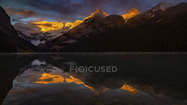 Luz solar dourada iluminando os picos da montanha e refletindo na água tranquila do Lago Louise, Banff National Park; Alberta, Canadá — Fotografia de Stock