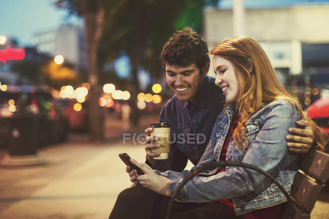 Молода пара сидить на лавці на вулиці в сутінках, дивлячись на смартфон — стокове фото