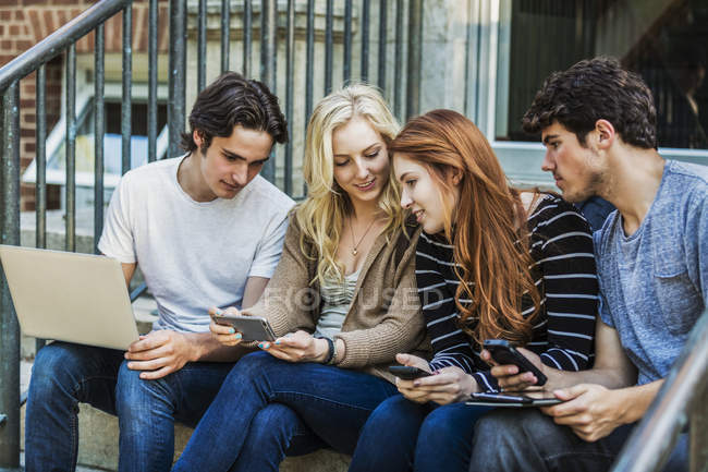 Vier Studenten, die in einer Reihe auf einer Treppe sitzen und ihre drahtlosen Geräte auf dem Universitätscampus benutzen — Stockfoto