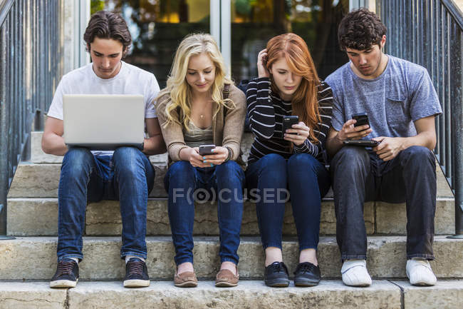 Quattro studenti seduti in fila su un gradino utilizzando i loro dispositivi wireless nel campus universitario — Foto stock