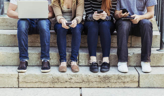 Quatre étudiants assis dans une rangée sur une marche en utilisant leurs appareils sans fil sur le campus de l'université — Photo de stock