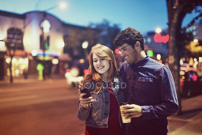 Young couple walking down a sidewalk on street at dusk while looking at a smartphone — Stock Photo